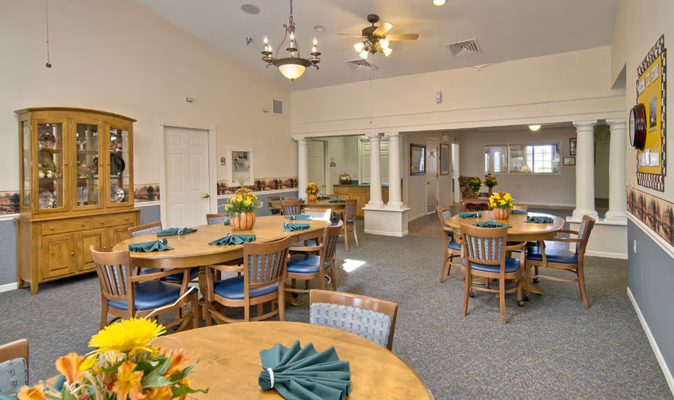 A dining room in a senior living facility with several round wooden tables and chairs arranged neatly. Each table is decorated with a small floral centerpiece and green folded napkins. The room features carpeted flooring, a wooden cabinet displaying decorative items, ceiling fans, and chandeliers. The walls are painted in light colors with a decorative border and columns separating the dining area from an adjacent hallway.