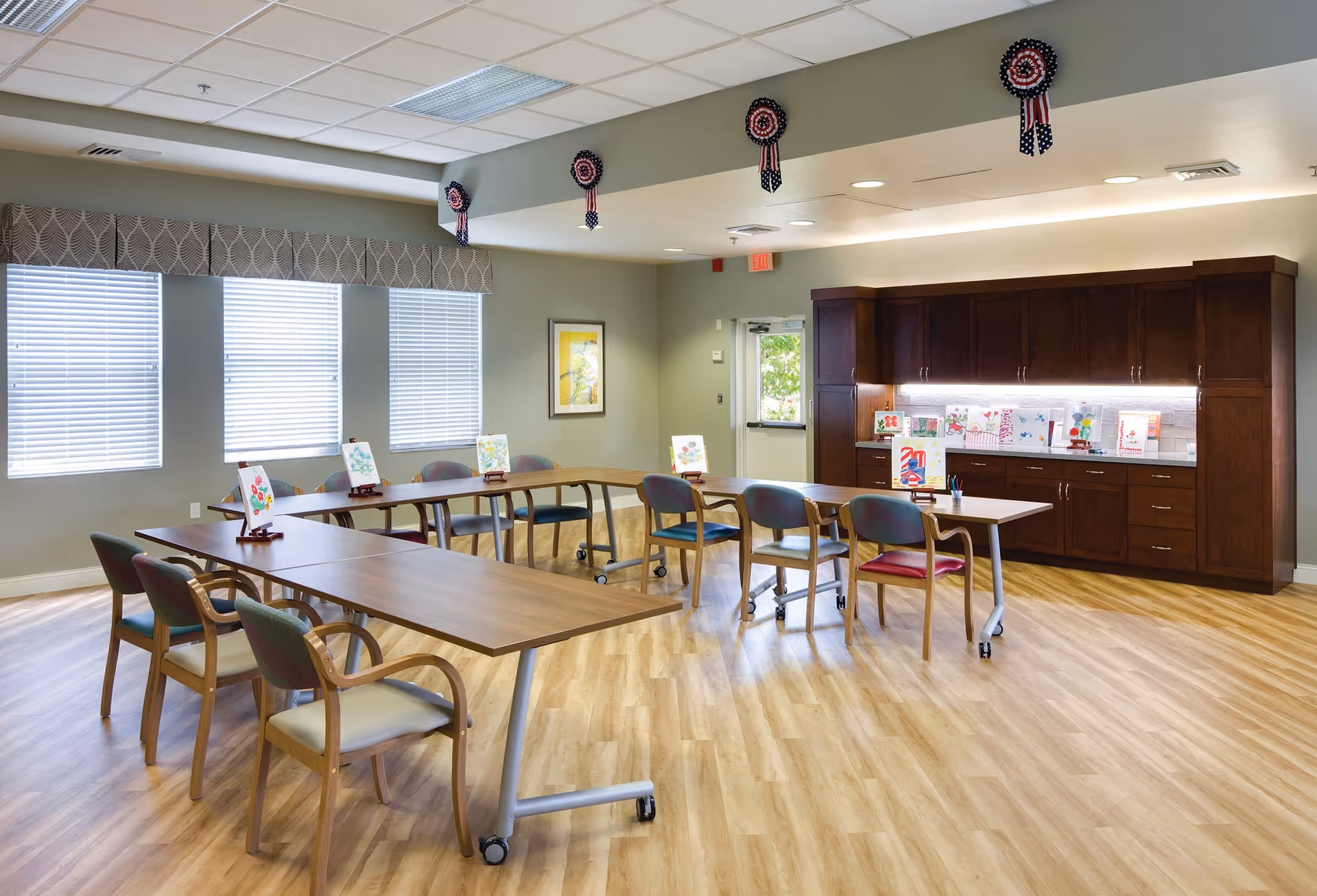 A bright activity room with wooden floors and light green walls featuring a U-shaped arrangement of tables and chairs. On the tables are small easels displaying colorful paintings. The back wall has dark wooden cabinets with more artwork displayed on the countertop. The ceiling has patriotic red, white, and blue rosette decorations hanging. Three large windows with blinds allow natural light into the room.