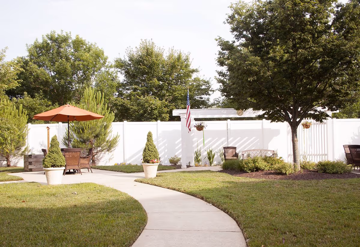 Outdoor garden area with a curved concrete pathway, green grass, potted plants, trees, and outdoor seating including chairs and a table with an orange umbrella. A white fence and an American flag are visible in the background.