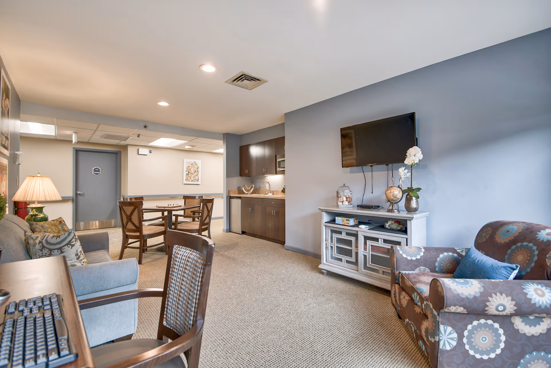 A cozy common area in a senior living facility featuring a patterned armchair with a blue cushion, a gray sofa with decorative pillows, a wooden table with chairs, a wall-mounted flat screen TV above a white cabinet with decorative items, and a kitchenette with dark cabinets and a countertop. The room has beige carpet and soft lighting.