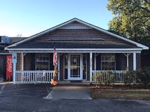 Single-story care facility with a front porch, white railing, and an American flag.