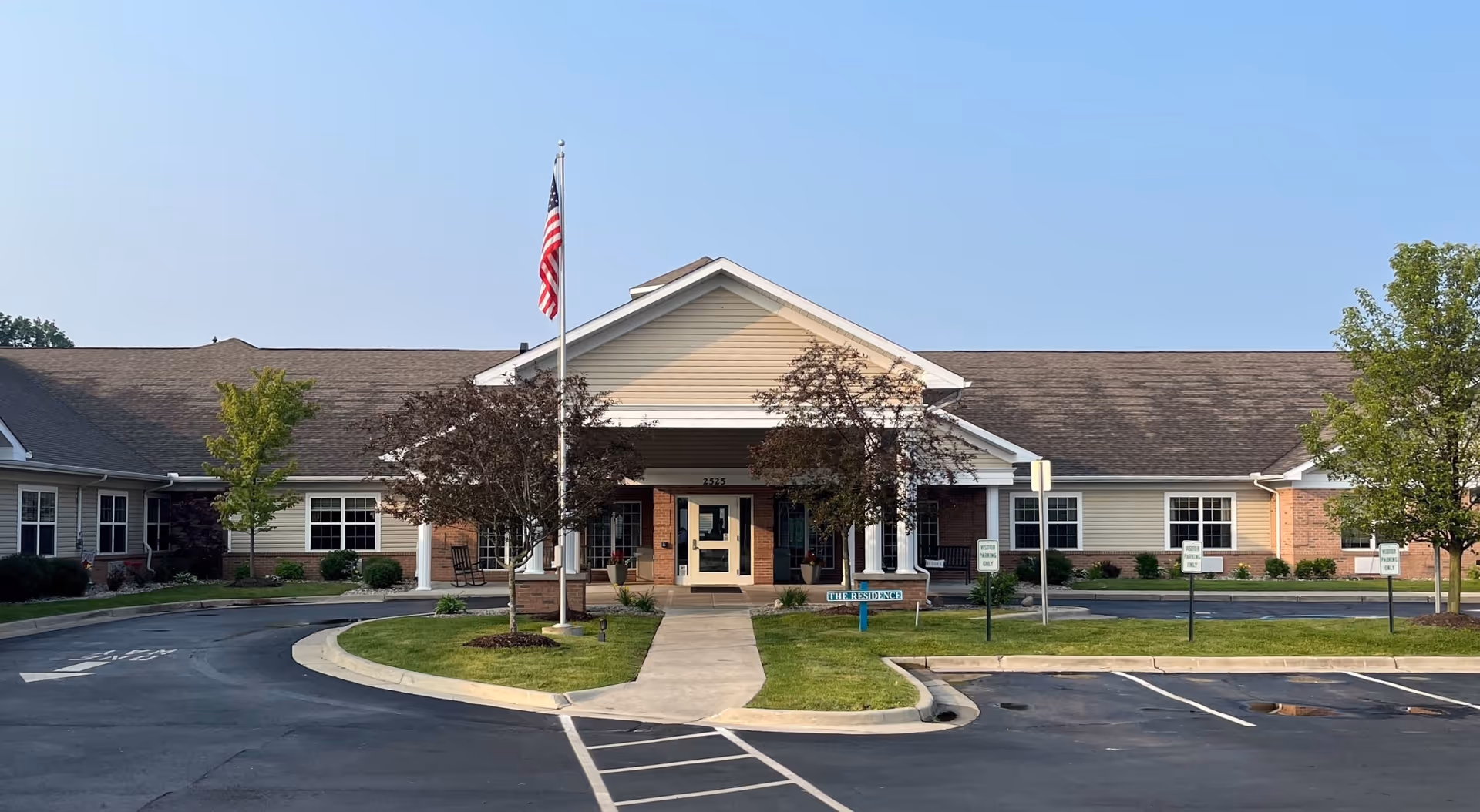 Front exterior view of Stonegate Health Campus building with a flagpole displaying the American flag, a paved driveway, and landscaped greenery including trees and bushes under a clear blue sky.