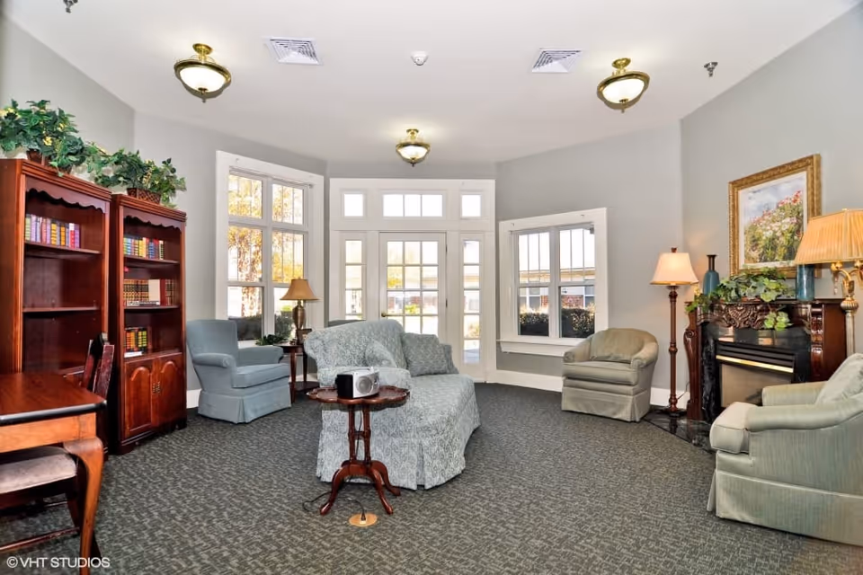 A cozy senior living facility common area with light blue upholstered armchairs and a loveseat arranged around a small wooden table. The room features large windows and glass doors letting in natural light, a wooden bookshelf filled with books, a fireplace with decorative plants and a framed painting above it, and several lamps providing warm lighting. The carpet is patterned and the walls are painted light gray.