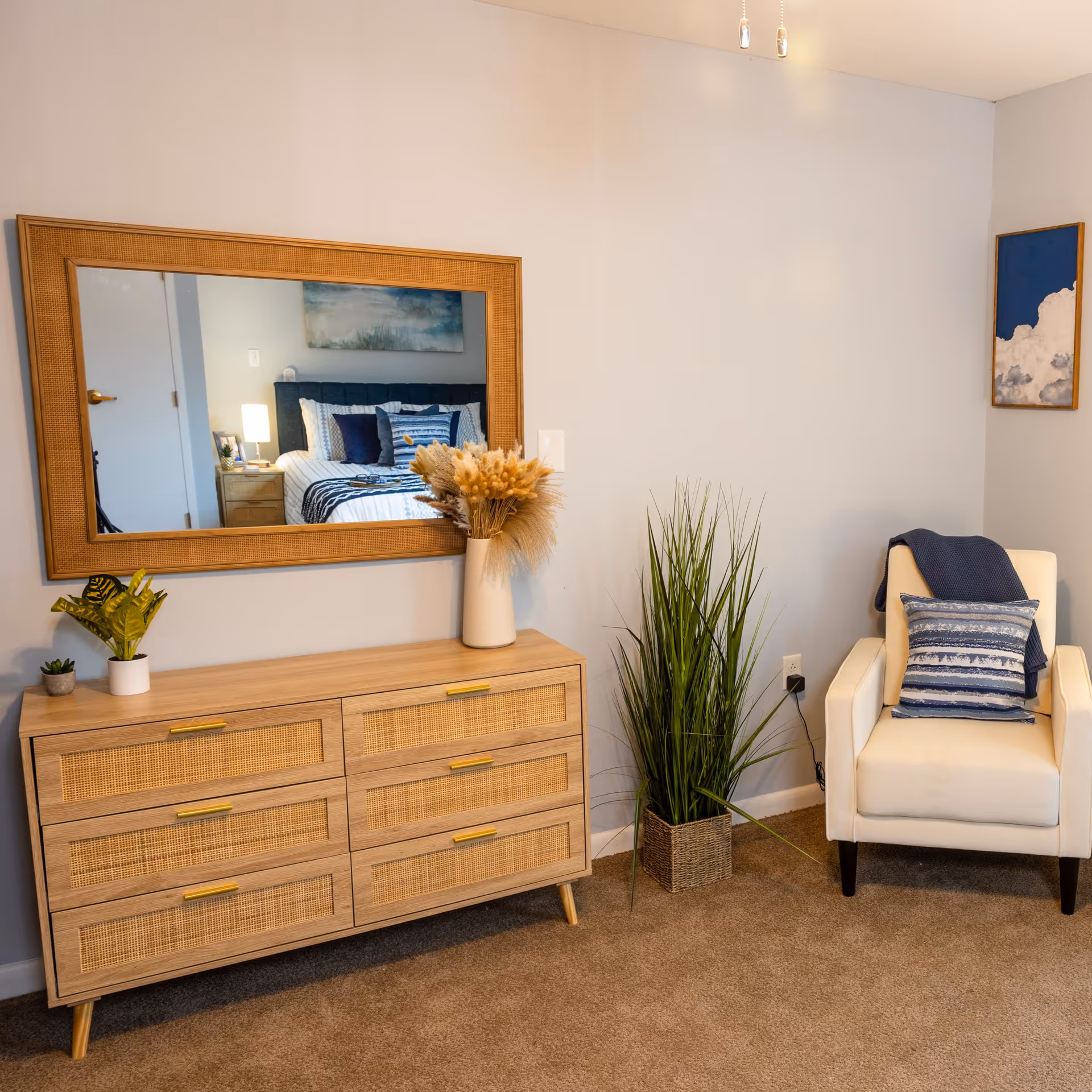 A cozy corner of a senior living bedroom featuring a wooden dresser with six drawers and a large rectangular mirror above it. On top of the dresser are three small potted plants and a tall vase with dried flowers. To the right of the dresser is a tall green plant in a woven basket. Next to the plant is a white armchair with a blue throw blanket draped over the back and a blue patterned pillow on the seat. The walls are light-colored with a framed artwork of clouds hanging above the chair. The carpet is beige.
