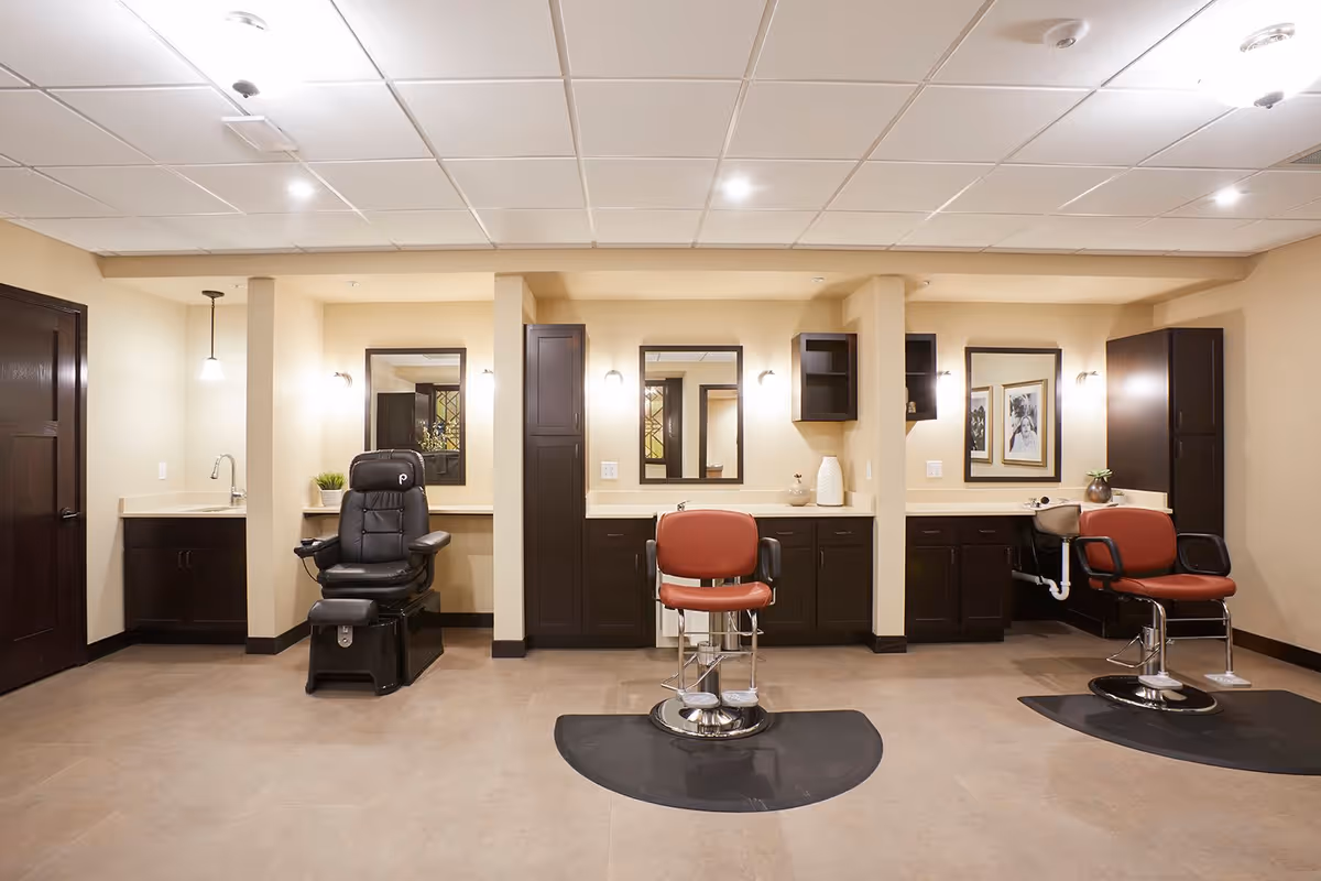 Interior view of a salon area in a senior living facility with three styling stations. Each station has a mirror, dark wood cabinetry, and a salon chair. The left station has a black salon chair, while the middle and right stations have red salon chairs. The room has beige walls, a tiled floor, and a white drop ceiling with recessed lighting.