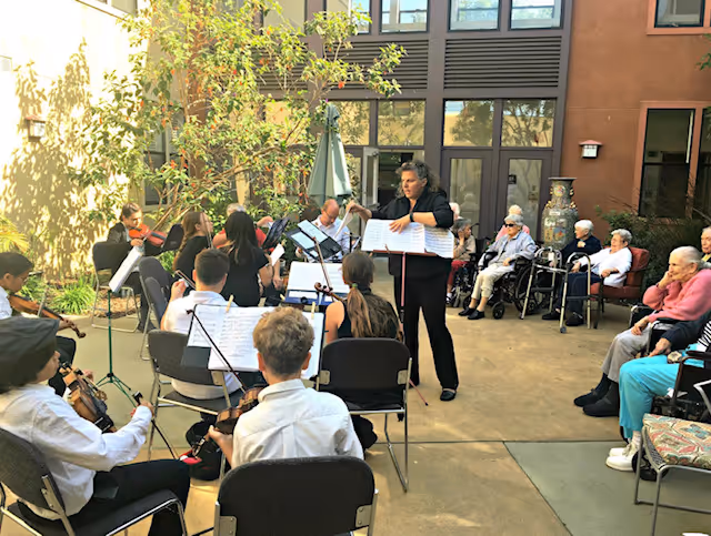 A group of young musicians playing string instruments in an outdoor courtyard of a senior living facility, with a conductor leading them. Several elderly residents are seated around the courtyard watching the performance.