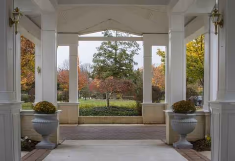 A covered entrance with white columns, large planters, and autumn trees visible beyond.
