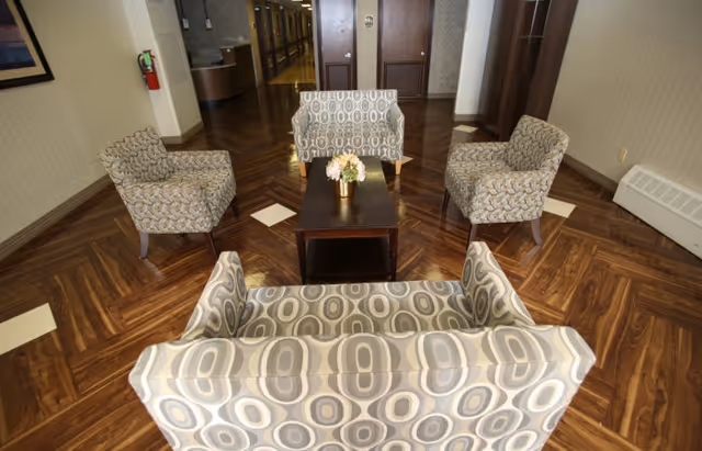 A seating area in a hallway with four patterned armchairs and a small wooden coffee table with a flower arrangement on top. The floor has a wood pattern with square white tiles, and there are doors and a fire extinguisher visible in the background.