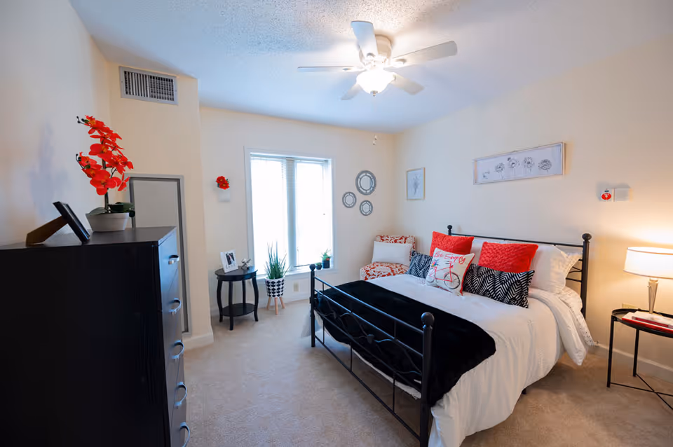 Well-lit bedroom with a black metal bed dressed in white bedding and red accent pillows, a dresser, side tables, plants, and a ceiling fan.