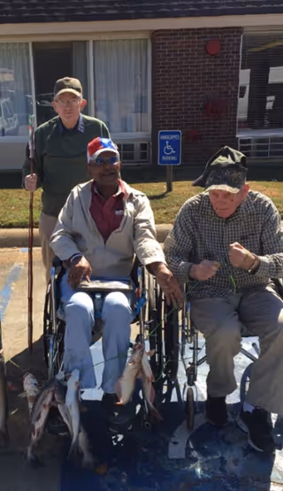 Three elderly men outside a building, two seated in wheelchairs and one standing behind them holding a fishing rod. The men in wheelchairs are holding several caught fish. There is a handicapped parking sign and a brick building with windows in the background.