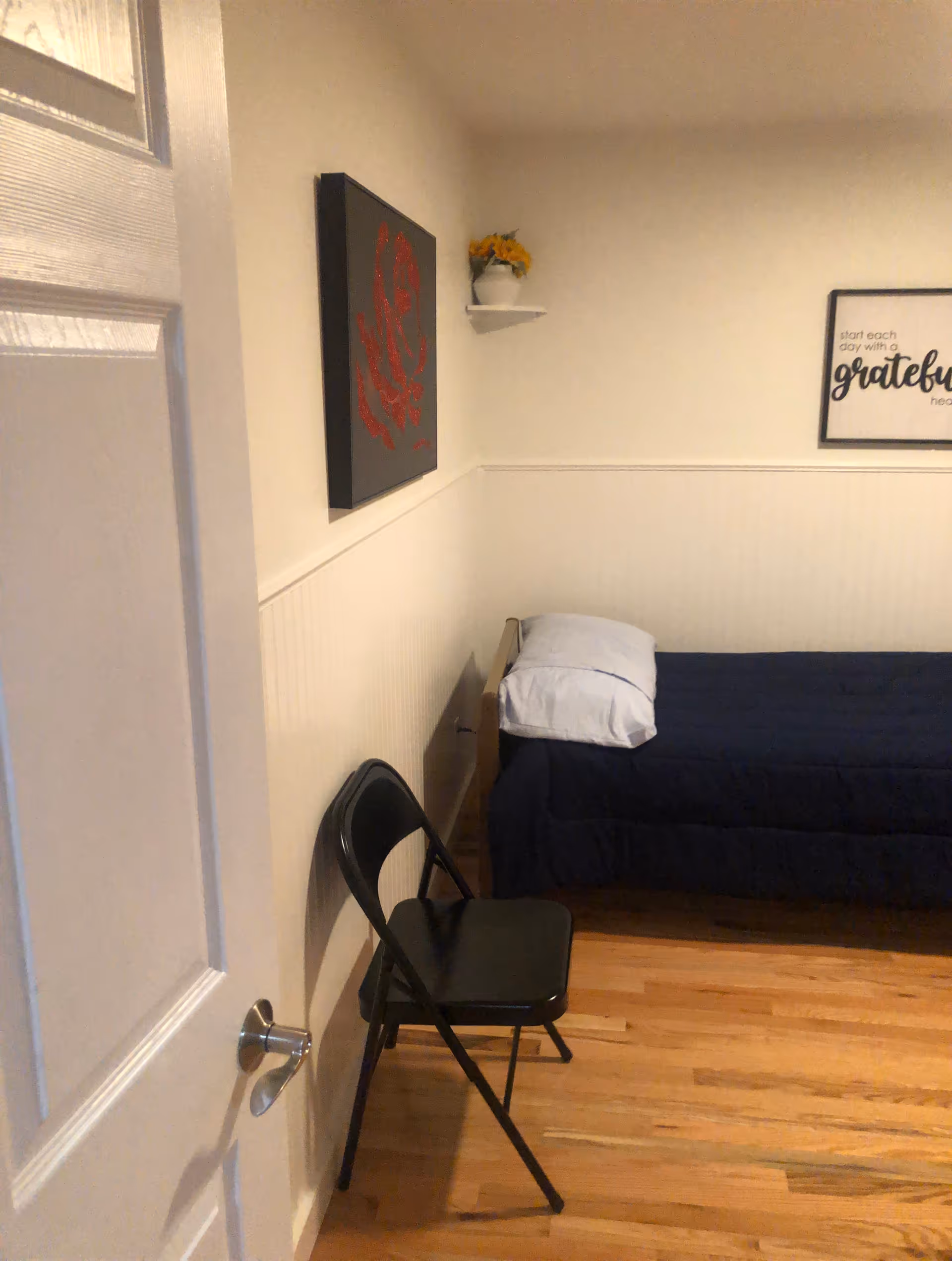 Small bedroom with a single bed against the wall, a black folding chair, and framed wall art.