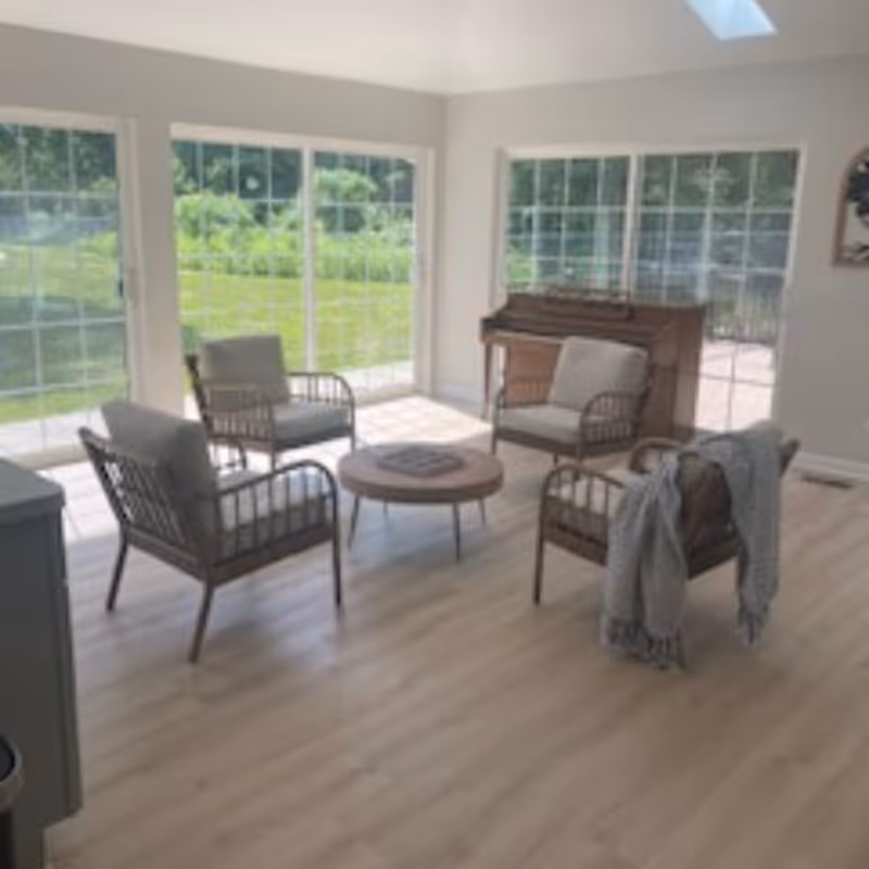 Bright sunroom-style living area with four cushioned wicker chairs around a round coffee table, large sliding glass doors, and a piano against the wall.