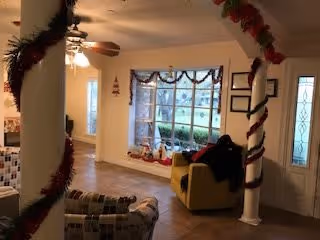 A cozy living room area decorated for the holidays with garlands wrapped around white columns and hanging from the ceiling. The room features a large window with a view outside, a yellow armchair, a patterned sofa, and a ceiling fan with lights. The floor is tiled, and there is a front door with decorative glass panels.