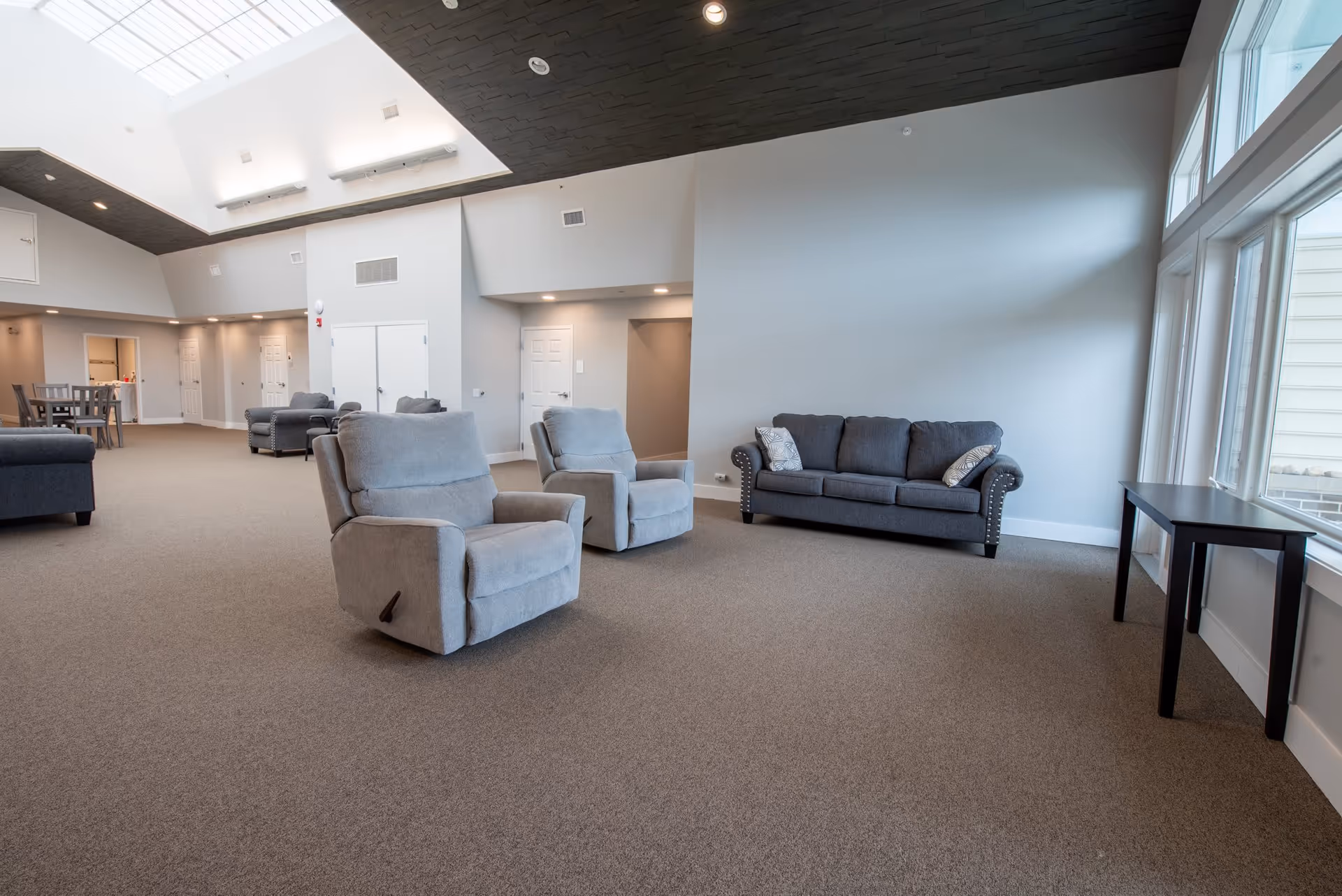 A spacious living room area with two light gray recliner chairs and a dark gray sofa with two patterned pillows. The room has large windows on the right side allowing natural light to enter, a high ceiling with a skylight, and neutral-colored walls and carpet. In the background, there is a dining table with chairs and multiple doors along the wall.