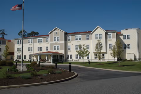 Exterior view of a three-story senior living facility building with beige and white siding, a red roof, and multiple windows. There is a driveway leading to a covered entrance, landscaped greenery, and an American flag on a flagpole in front of the building.