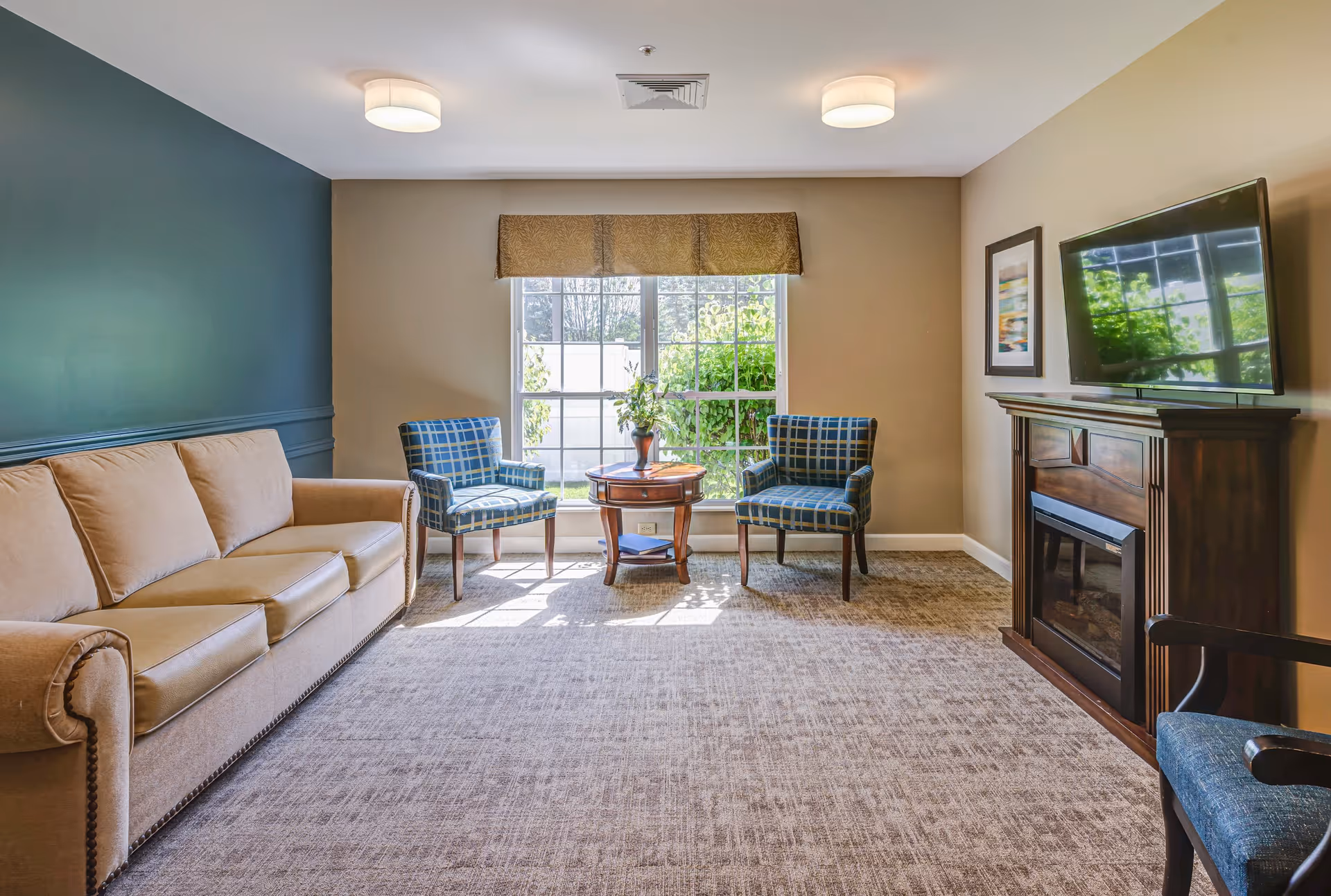 A cozy living room with a beige sofa on the left, two blue plaid armchairs around a small wooden table with a vase of flowers in front of a large window with a brown valance. On the right side, there is a wooden electric fireplace with a flat-screen TV mounted above it and a framed picture on the wall. The room has beige walls, a dark green accent wall, carpeted floor, and two ceiling lights.