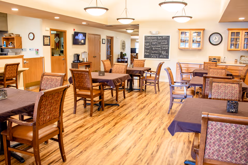A dining room in a senior living facility with multiple wooden tables covered with dark tablecloths and matching wooden chairs with patterned upholstery. The room has wood flooring, warm lighting from ceiling fixtures, a chalkboard menu on the wall, a clock, and cabinets with glass doors. There is a small counter area with a coffee machine and a TV mounted on the wall.