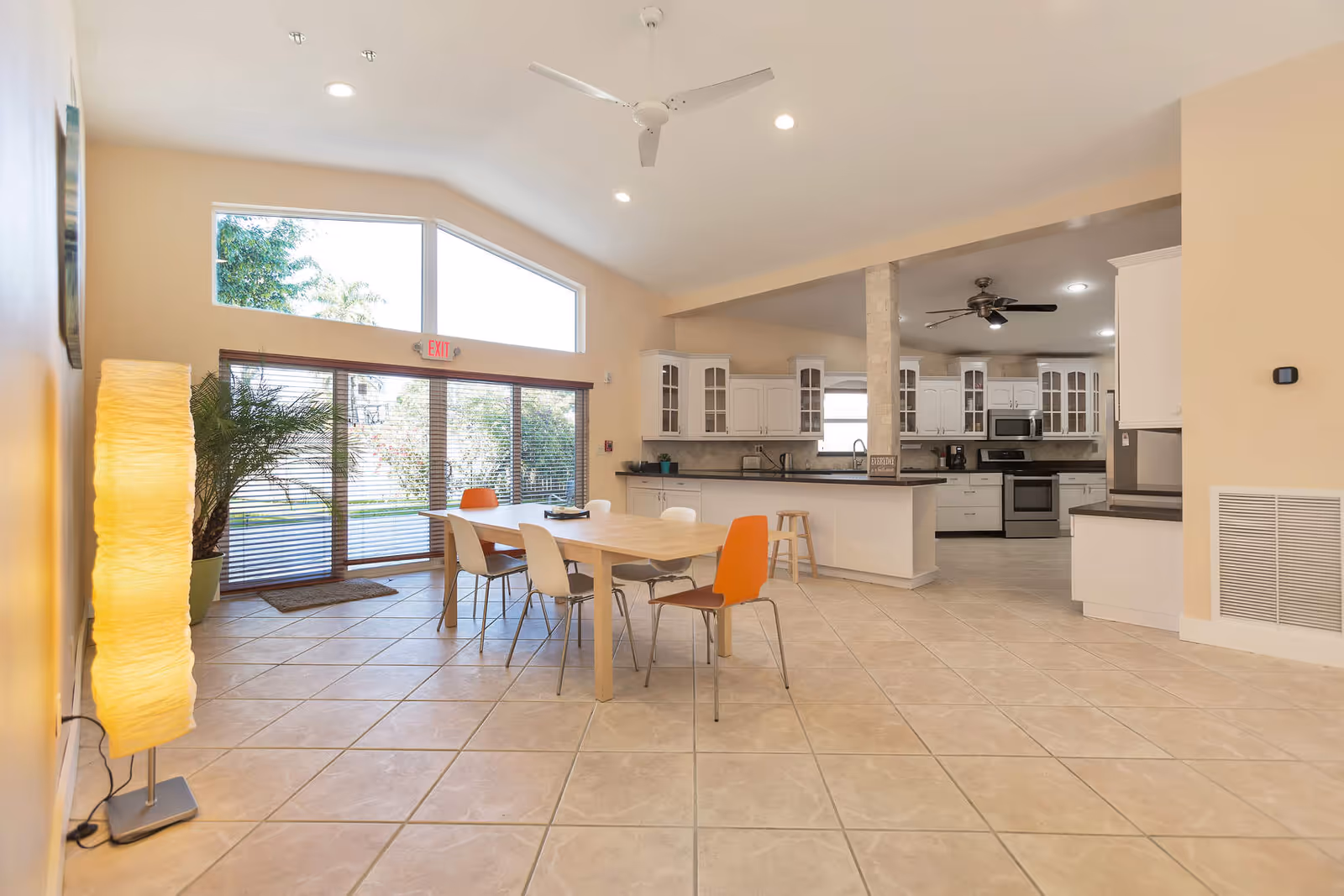 Bright and spacious interior of an assisted living facility showing a dining area with a wooden table and six chairs, some white and some orange. Large windows with blinds let in natural light and offer a view of greenery outside. The room has beige tiled floors, a tall floor lamp, and a ceiling fan. In the background, there is a modern kitchen with white cabinets, stainless steel appliances, and a countertop with stools.