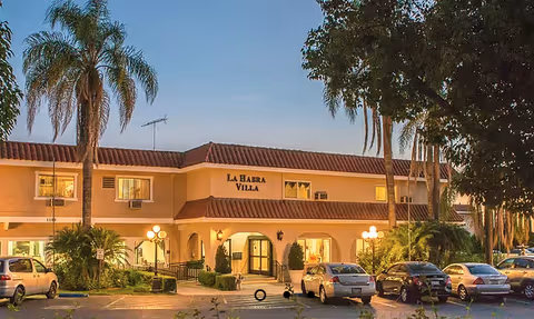 Exterior view of La Habra Villa, a two-story retirement facility with a tiled roof, palm trees, and several parked cars in front during dusk.