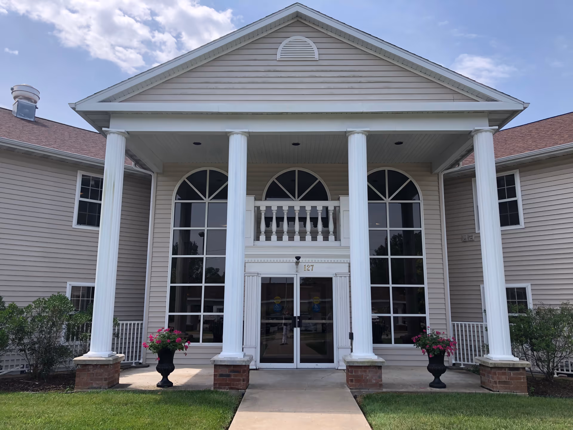 Front entrance of a retirement community building with tall white columns, arched windows, double glass doors and potted flowers.