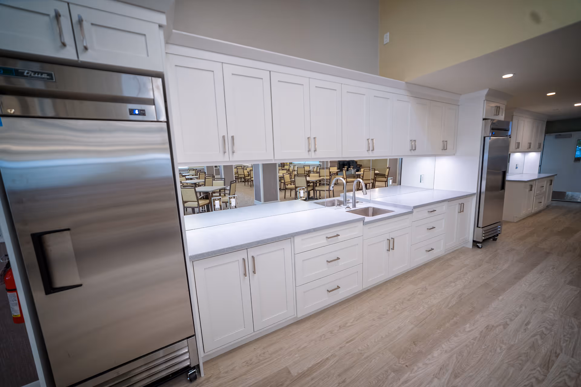 A modern kitchen area with white cabinets, stainless steel refrigerators, a double sink, and light-colored wooden flooring. In the background, a dining area with tables and chairs is visible through a large window-like opening in the wall.