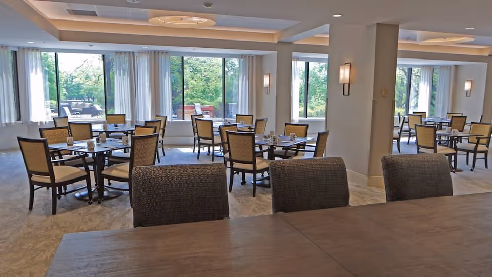 Bright communal dining room with multiple tables and chairs and large windows overlooking greenery.