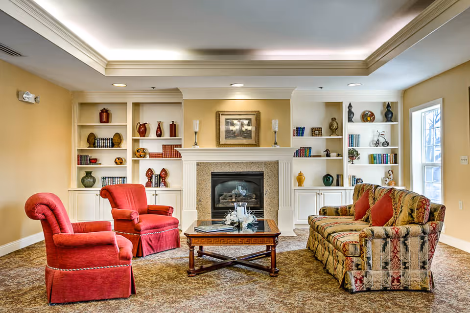 A cozy living room in a retirement community featuring two red upholstered armchairs and a patterned sofa with red cushions arranged around a wooden coffee table. The room has built-in white bookshelves filled with decorative items and books, and a fireplace with a framed picture above it. Natural light comes in through a window on the right.