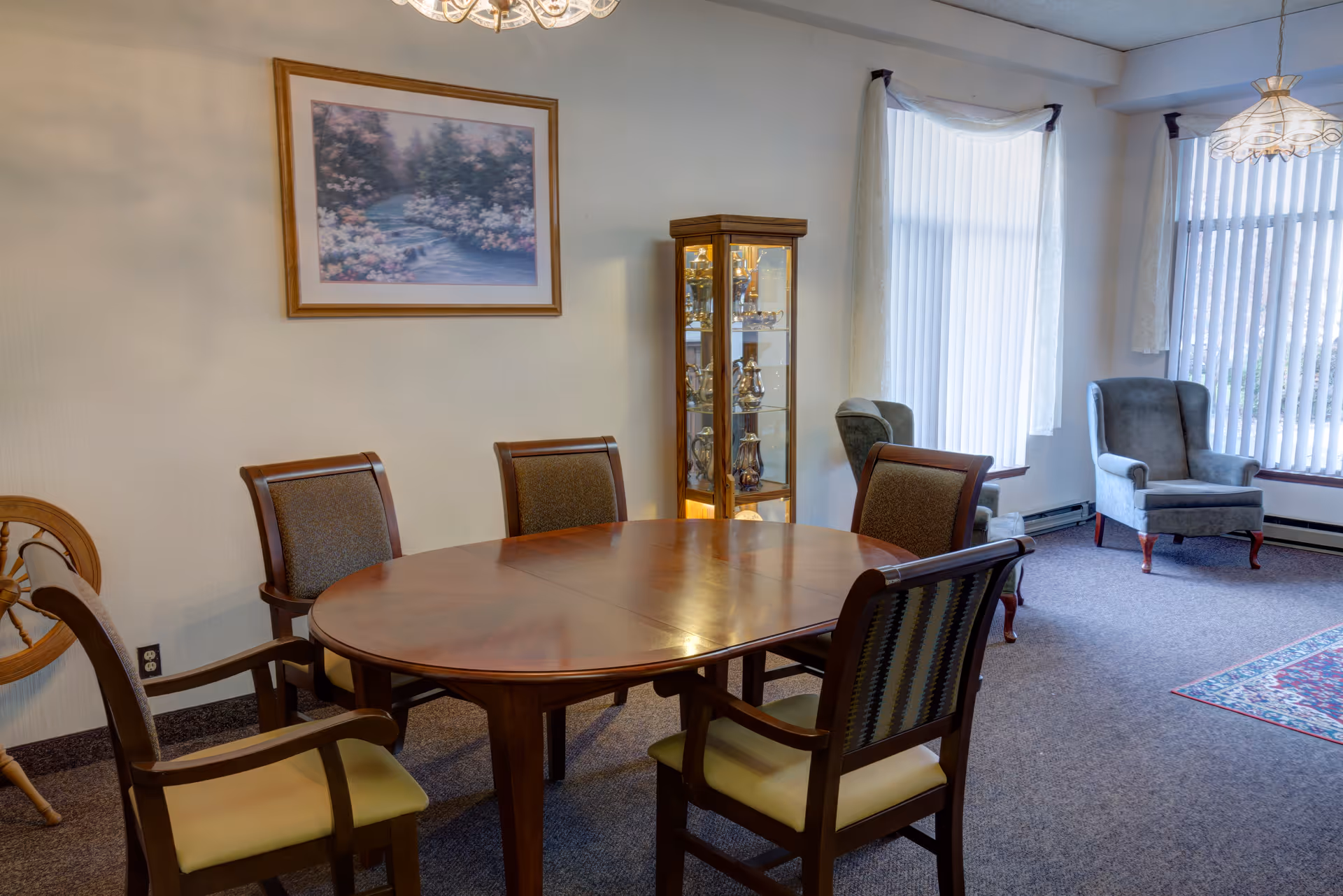 A dining area with a wooden oval table surrounded by six chairs with cushioned seats and backs. In the background, there is a glass display cabinet with decorative items inside, two upholstered armchairs near large windows with vertical blinds, and a framed painting of a floral landscape on the wall.