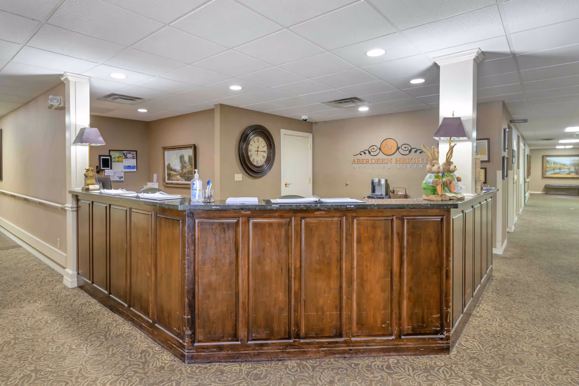Reception desk area inside Aberdeen Heights Assisted Living facility with a wooden counter, two decorative lamps, hand sanitizer, paperwork, a wall clock, framed paintings, and a logo on the wall behind the desk. A hallway extends to the right with additional framed pictures on the walls.