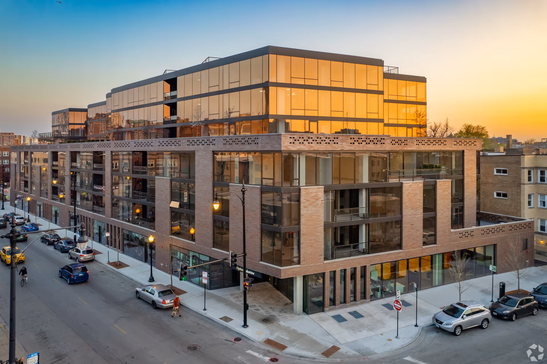 Modern multi-story building with large glass windows reflecting the sunset, situated on a city street corner with parked cars and a few pedestrians.