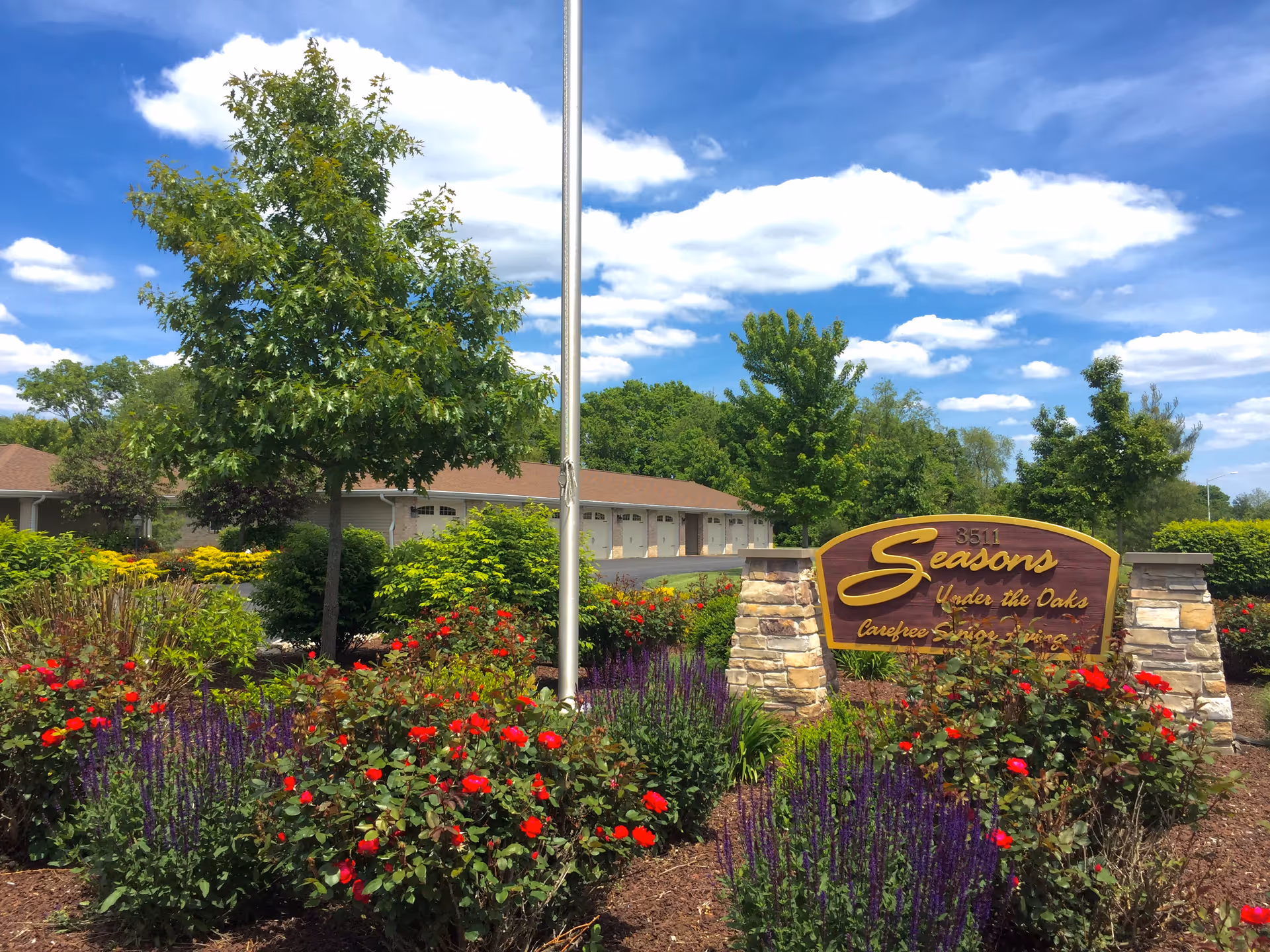 A landscaped garden area with vibrant red and purple flowers, green shrubs, and trees under a blue sky with scattered clouds. A stone and wood sign reads '3511 Seasons Under the Oaks Carefree Senior Living' in the foreground. A building with multiple garage doors is visible in the background.