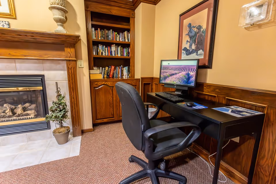 A cozy office corner in an assisted living facility featuring a black office chair and desk with a computer monitor displaying a lavender field. Behind the desk is a framed picture on the wall. To the left, there is a wooden built-in bookshelf filled with books and a fireplace with a small potted plant on the tiled hearth. The room has beige walls and carpeted flooring.