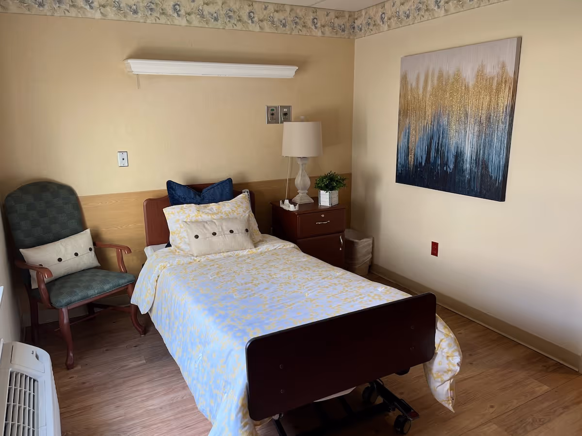 A single bed with a wooden headboard and footboard, covered with a white and yellow patterned bedspread and several pillows, is placed against a beige wall with floral wallpaper border near the ceiling. Next to the bed is a wooden nightstand with a lamp and a small potted plant. To the left of the bed is a green upholstered armchair with a decorative pillow. A large abstract painting with gold, blue, and white colors hangs on the adjacent wall. The floor is wooden, and there is an air conditioning unit near the bottom left corner.
