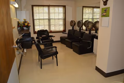 Small salon room with black styling chairs, hooded hair dryers, sinks and large windows.