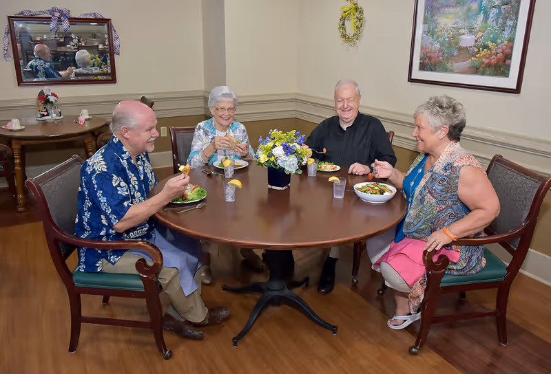 Four elderly residents sit around a round dining table smiling and eating salads in a dining room.