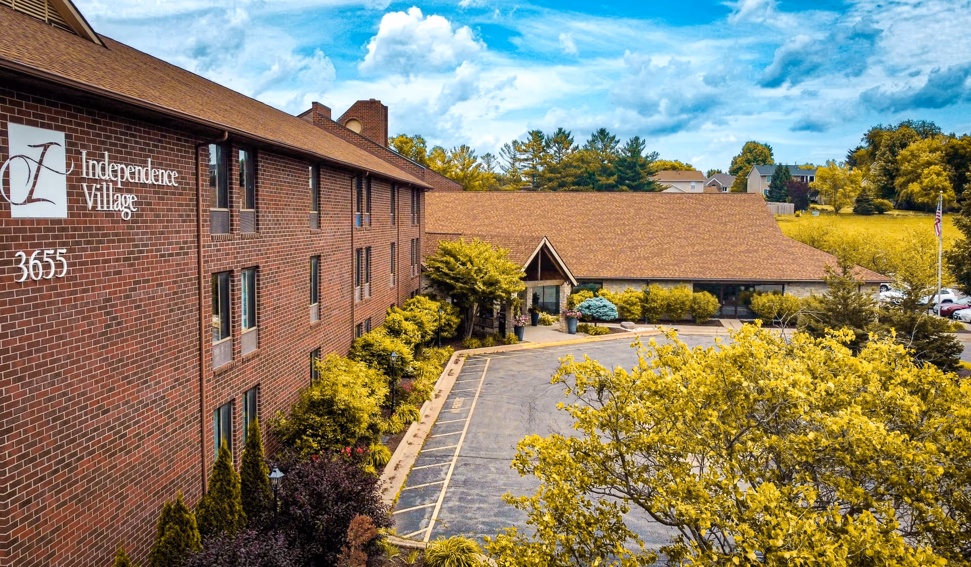 Exterior view of Independence Village, a multi-story brick building with a brown roof, surrounded by trees and greenery under a partly cloudy blue sky. The building has several windows and a driveway leading to the entrance.