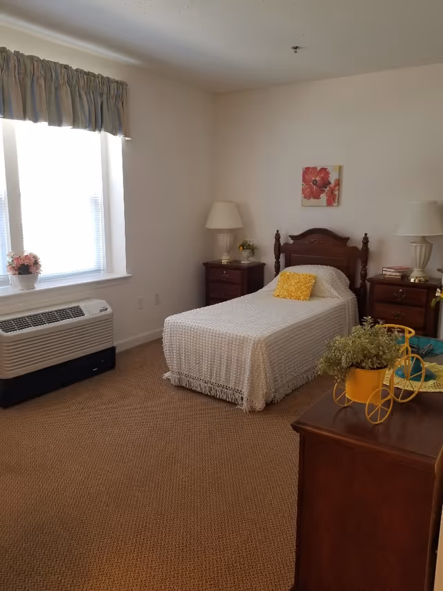 Well-lit single bedroom with a twin bed, two nightstands and lamps, a window with an air conditioner, and floral decor.