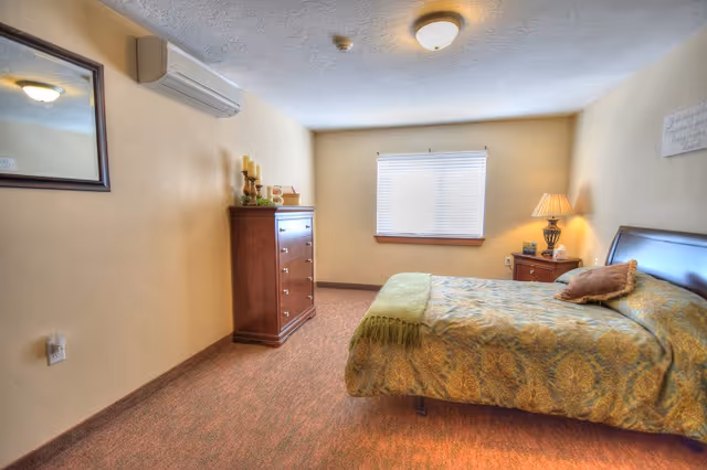 A cozy bedroom with beige walls and carpeted floor. The room features a bed with a patterned green and gold bedspread and a brown pillow. There is a wooden nightstand with a lamp and some small items on it next to the bed. Across from the bed is a tall wooden dresser with decorative candles and a letter B on top. A window with blinds is centered on the far wall, and a mirror hangs on the left wall. A ceiling light fixture and a wall-mounted air conditioning unit are also visible.