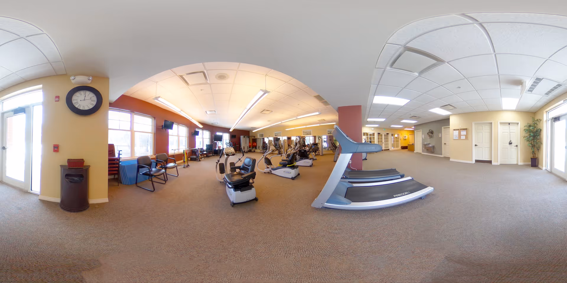 Wide panoramic view of a fitness room in a senior living facility with exercise equipment including treadmills, stationary bikes, and weight machines. The room has large windows letting in natural light, beige walls, and a clock on the wall near the entrance door. Chairs and exercise mats are placed along the windows, and the ceiling has fluorescent lighting panels.