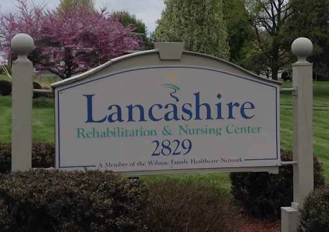 Outdoor sign for Lancashire Rehabilitation & Nursing Center with the address number 2829, surrounded by green grass, bushes, and trees with some purple blossoms in the background.