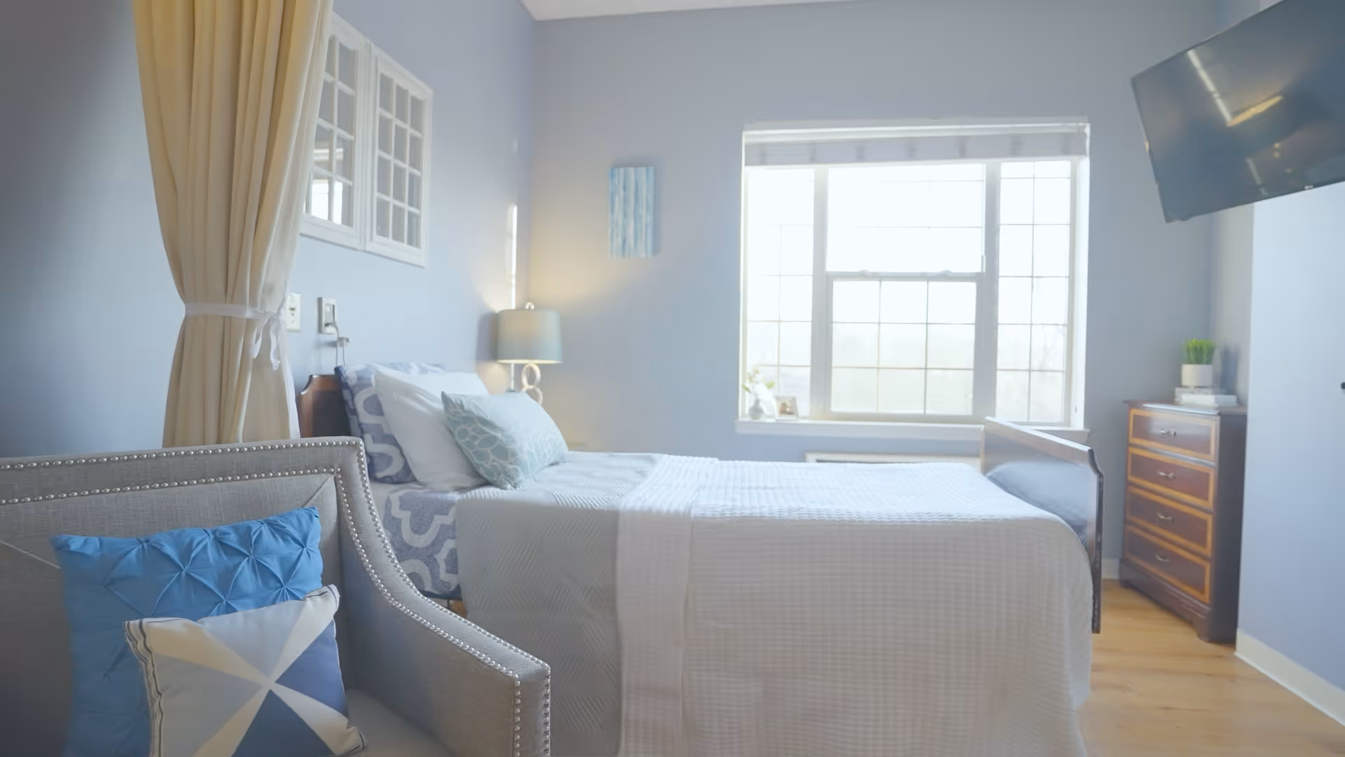 A bright and cozy bedroom with a single bed made up with white and patterned blue bedding. There is a gray armchair with blue and white pillows in the foreground, a window letting in natural light, a wooden dresser with books and a small plant, a wall-mounted TV, and light blue walls with minimal decor.