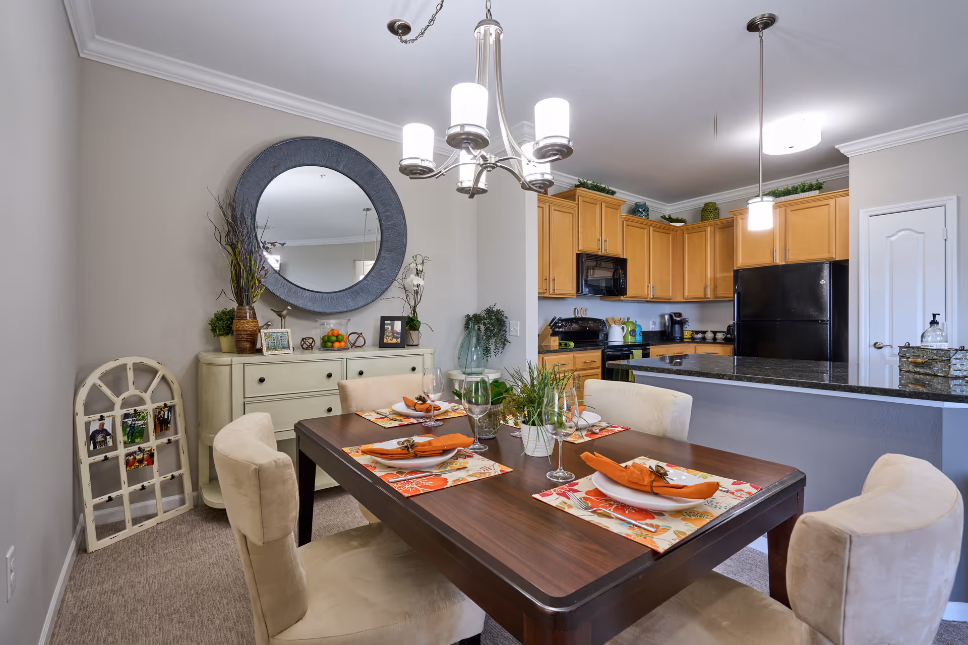 Dining area with a set table and upholstered chairs in front of an open kitchen with light wood cabinets and black appliances.