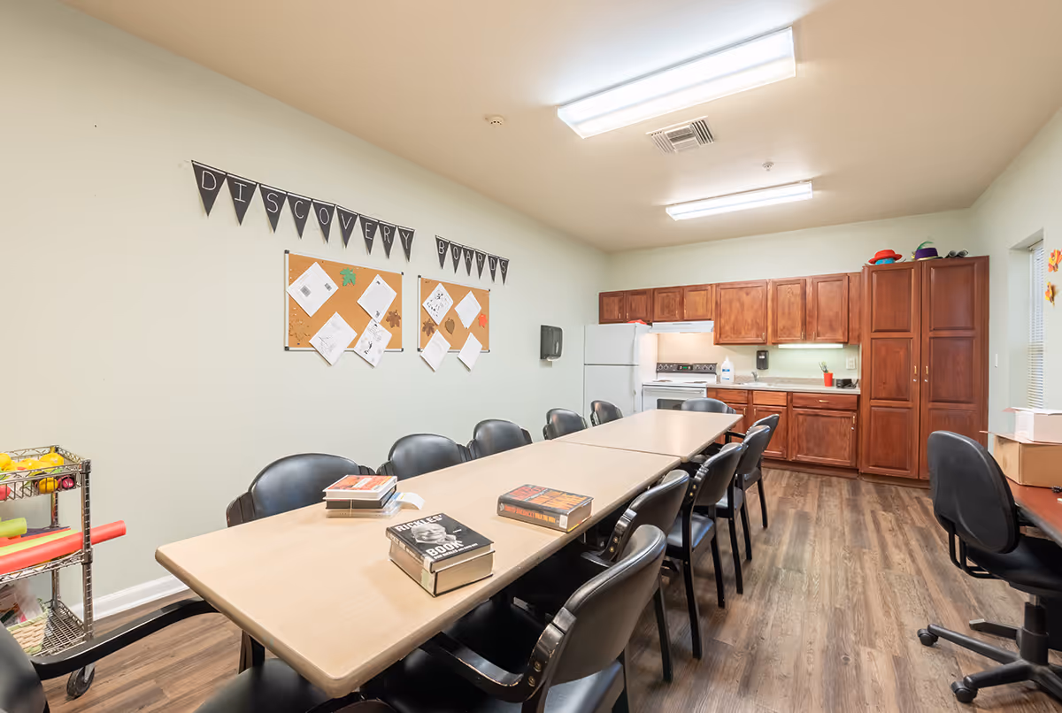 A multipurpose room with a long table surrounded by black chairs, several books on the table, a kitchen area with wooden cabinets, a refrigerator, stove, and sink in the background. On the wall, there are bulletin boards with papers and a banner that reads 'DISCOVERY BOARDS'. The floor is wood, and the room is brightly lit with ceiling lights.