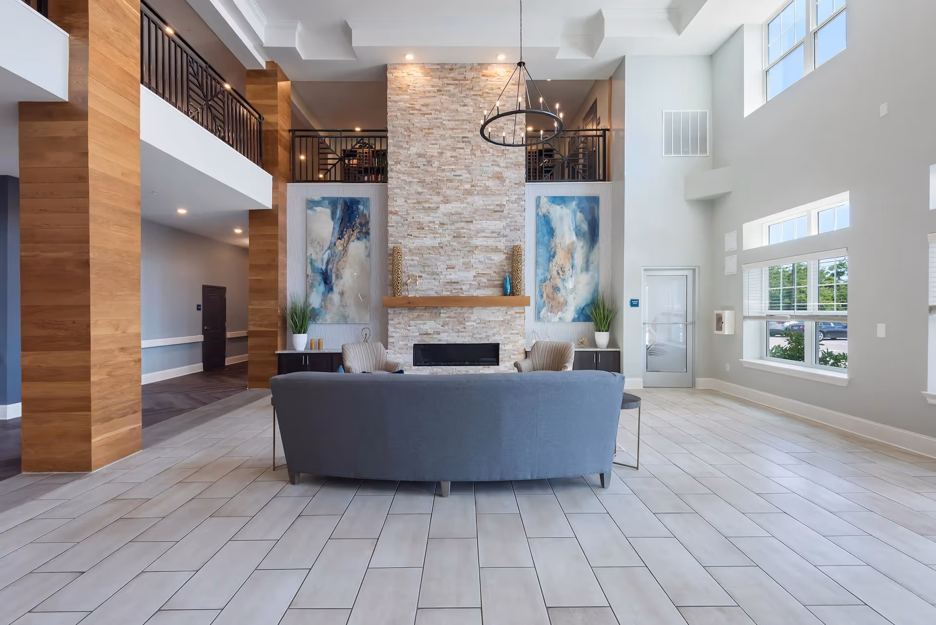 A spacious and modern living area with a high ceiling, large windows letting in natural light, a stone fireplace centered on the far wall, two abstract paintings on either side of the fireplace, a gray sofa facing the fireplace, and two beige armchairs. The room features light-colored tiled flooring and wooden columns, with a balcony railing visible on the upper level.