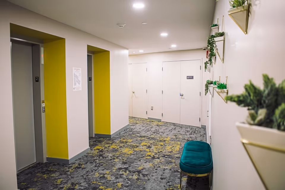 Interior hallway with two elevators on the left side, each framed by yellow walls. The floor is covered with a patterned carpet in shades of gray and yellow. On the right wall, there are decorative wall-mounted planters with green plants and a teal cushioned bench below them. At the end of the hallway, there are two closed white doors.