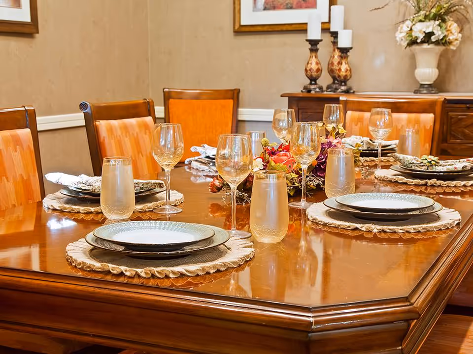 A polished wooden dining table set for six with plates, glasses, and cloth napkins on woven placemats. The chairs have orange patterned upholstery. In the background, there is a wooden sideboard with decorative candles and a floral arrangement.