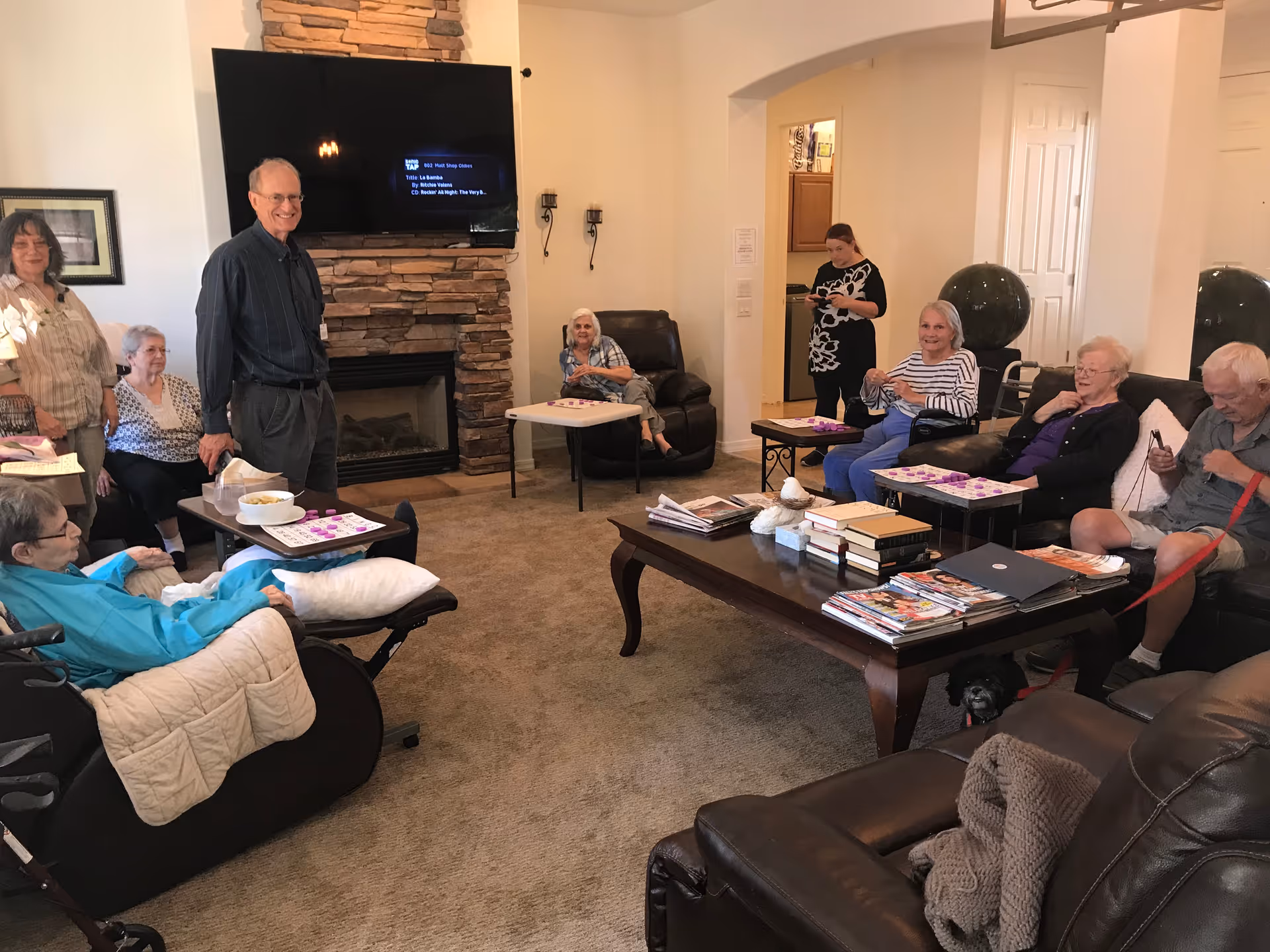 A group of seniors gathered in a comfortable living room with sofas, a fireplace and a coffee table, playing games.