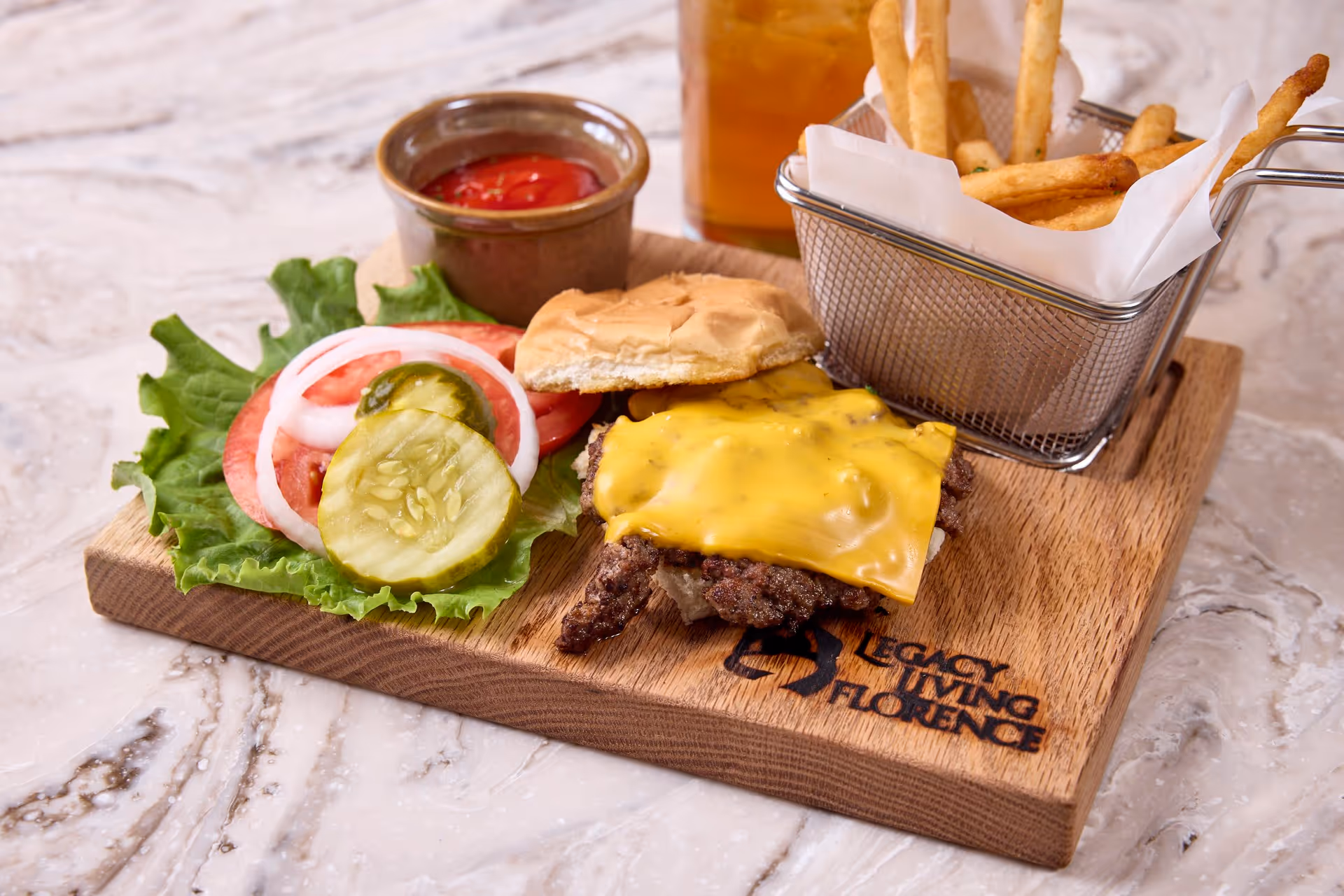 A cheeseburger with melted cheddar cheese on a bun, served open-faced with lettuce, tomato, onion, and pickles on the side. Next to the burger is a small metal basket filled with French fries and a small cup of ketchup on a wooden board engraved with the text 'Legacy Living of Florence'.