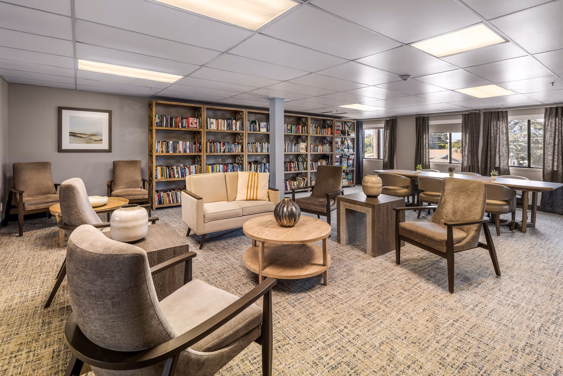 A spacious senior living community lounge area with comfortable armchairs and a sofa arranged around wooden coffee tables. The room features large bookshelves filled with books along one wall, large windows with curtains letting in natural light, and a long table with chairs near the windows. The decor is warm and inviting with neutral tones and decorative vases on the tables.