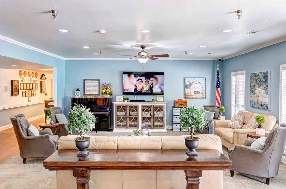 Bright communal living room with sofas, armchairs, a TV and piano, decorated with plants and artwork.